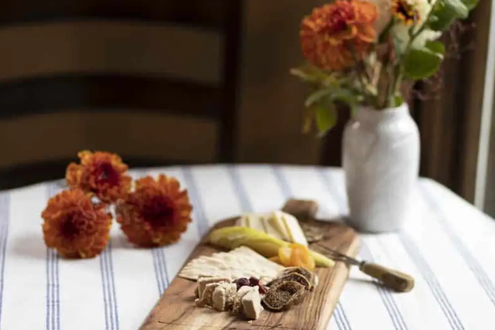 A wooden board with assorted cheeses, dried fruits, and nuts, accompanied by a vase of orange flowers on a striped tablecloth.