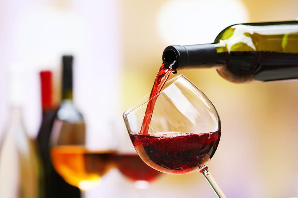 A wine bottle pours red wine into a glass, with other wine bottles blurred in the background.