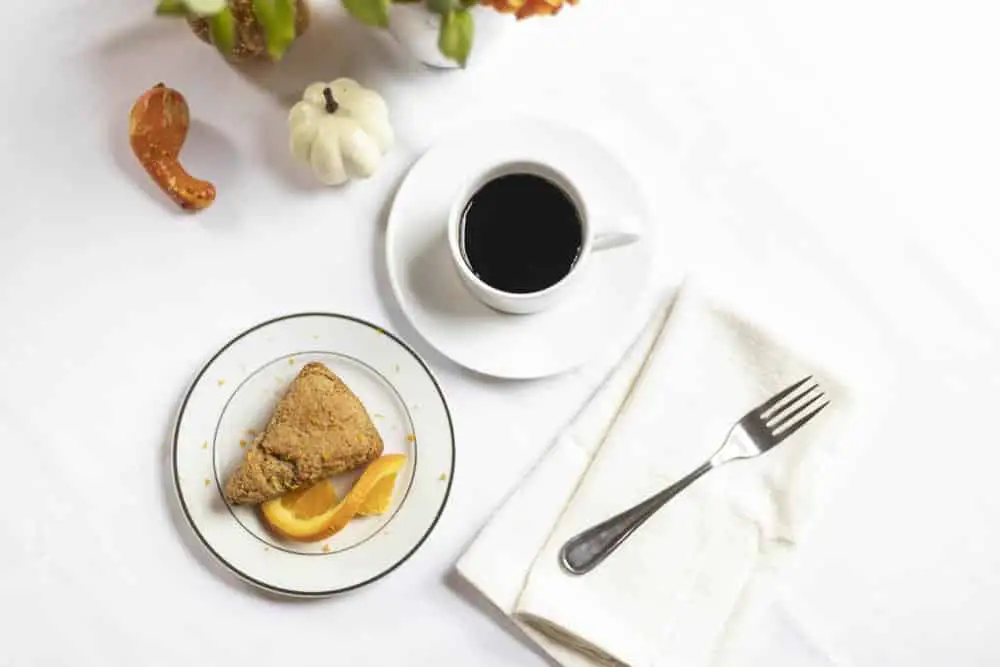 A plate of scone and orange slices beside a cup of coffee on a white tablecloth, with a fork and decorative elements.