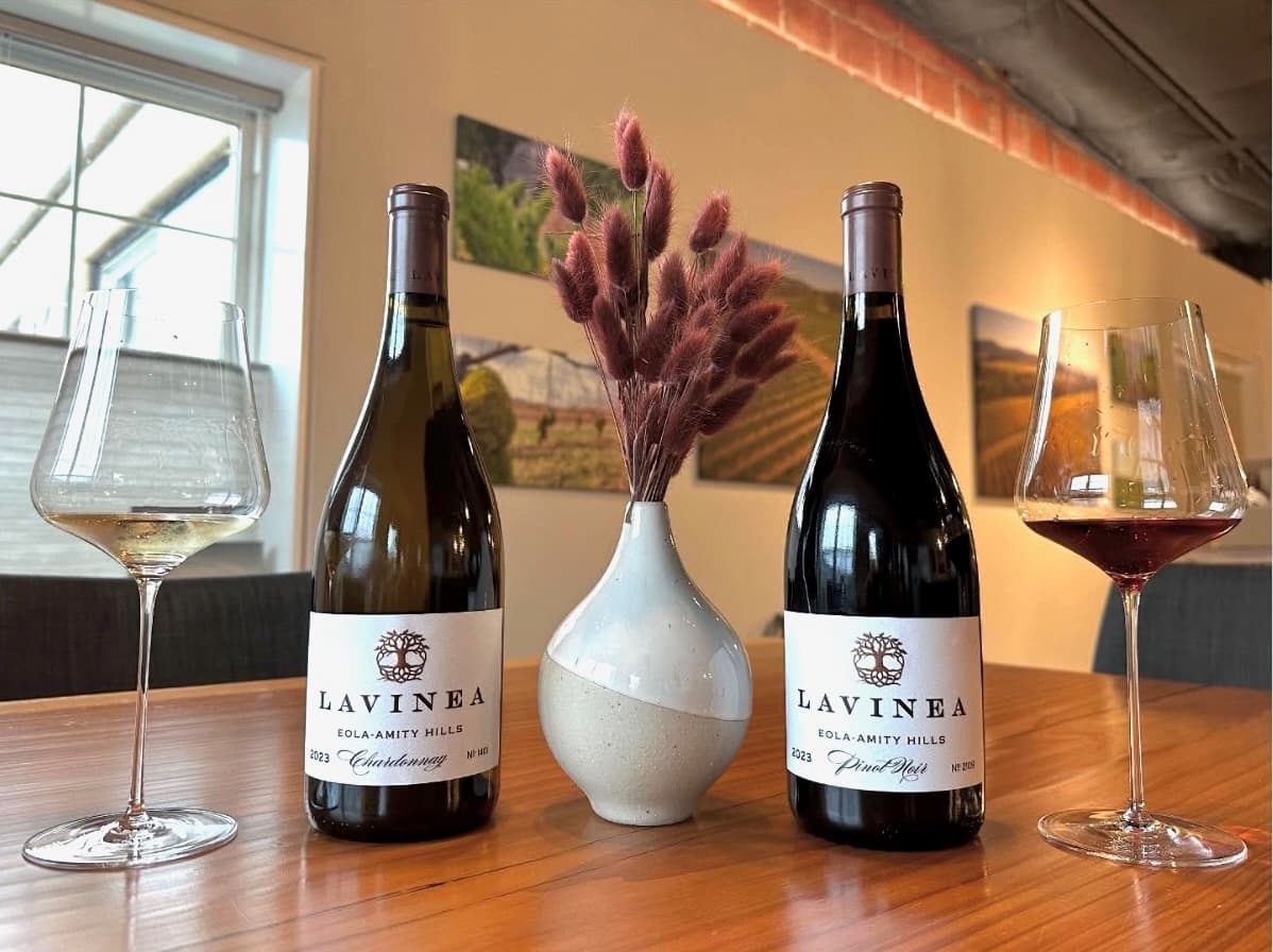 Two bottlesw of Lavinea wine sit on a wooden table with 2 wine glasses beside them and a floral arrangement in between