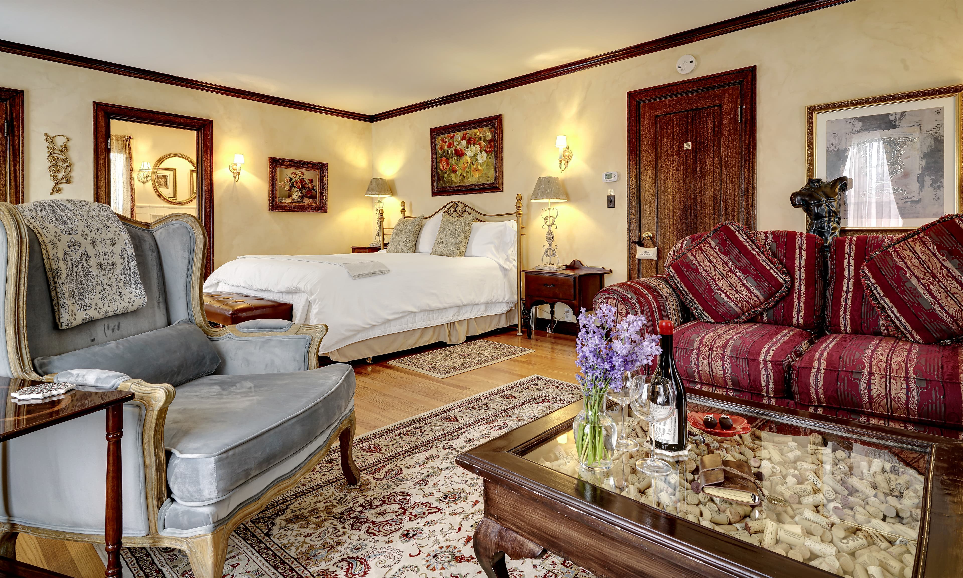 A cozy hotel room featuring a bed, elegant armchairs, a wine bottle, and decorative artwork.