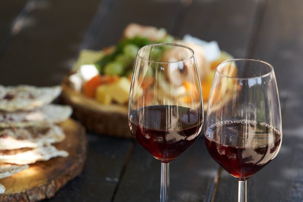 Two glasses of red wine in the foreground with a colorful spread of snacks in the background.