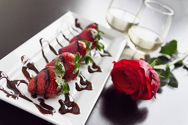 A plate of chocolate-drizzled strawberries accompanied by a red rose and two glasses of wine.