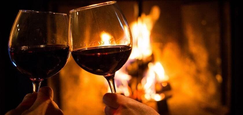 Two glasses of red wine clink together in front of a cozy fireplace.
