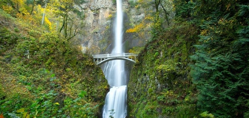 A majestic waterfall cascades down a rocky cliff, with a bridge spanning the gorge below, surrounded by autumn foliage.