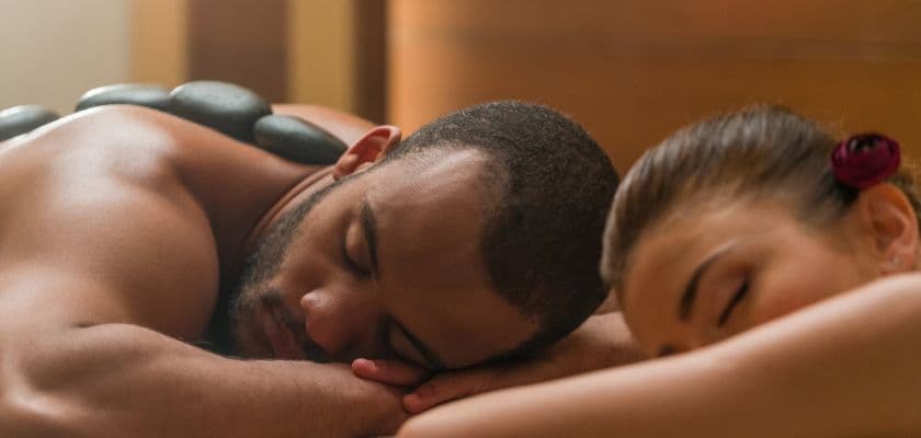 A man and a woman are peacefully receiving a massage side by side with hot stones on their backs.