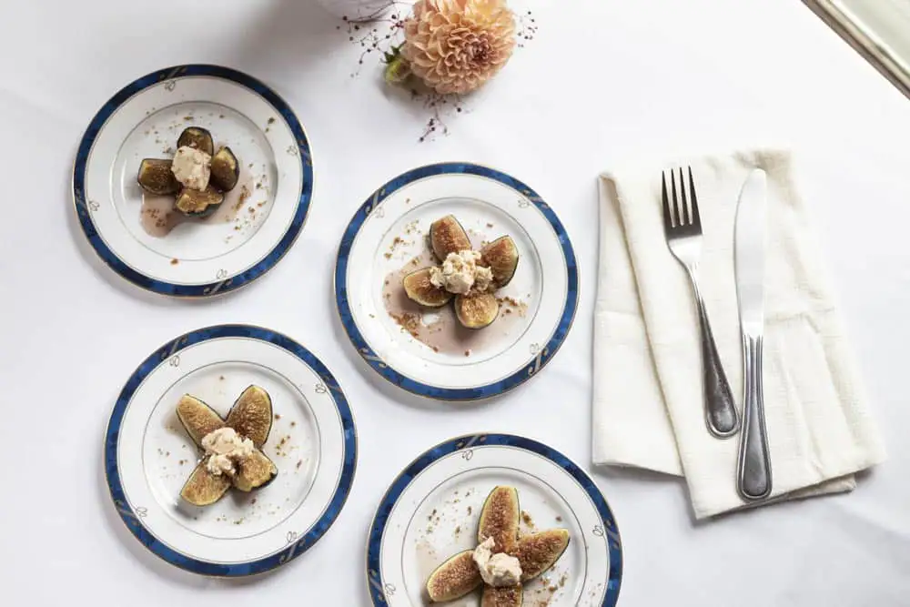 Four plates of elegantly arranged figs topped with cream, accompanied by a fork and knife on a neatly set table.