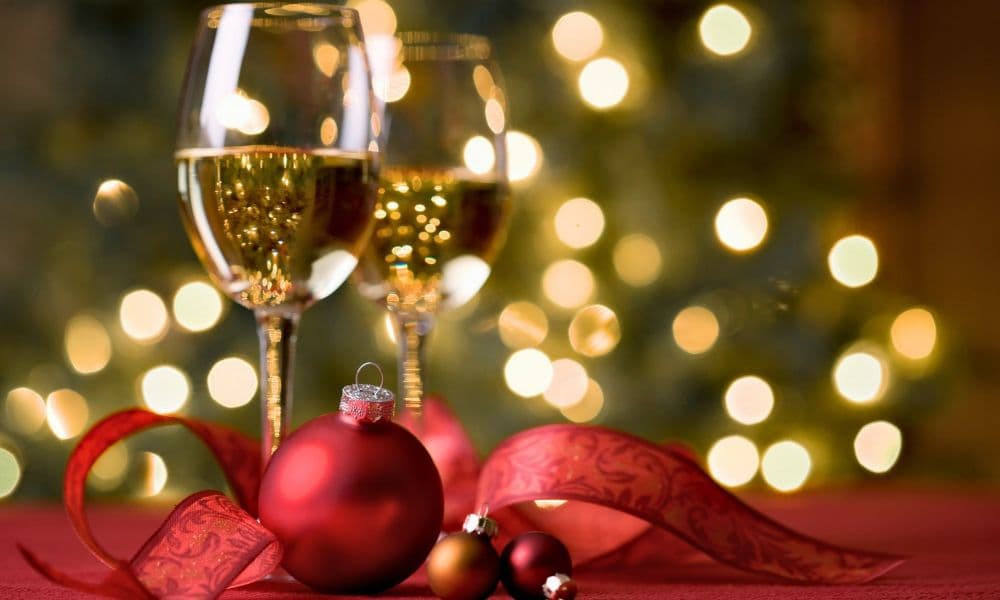 Two glasses of sparkling white wine next to a red Christmas bulb and ribbon with holiday lights in the backdrop.