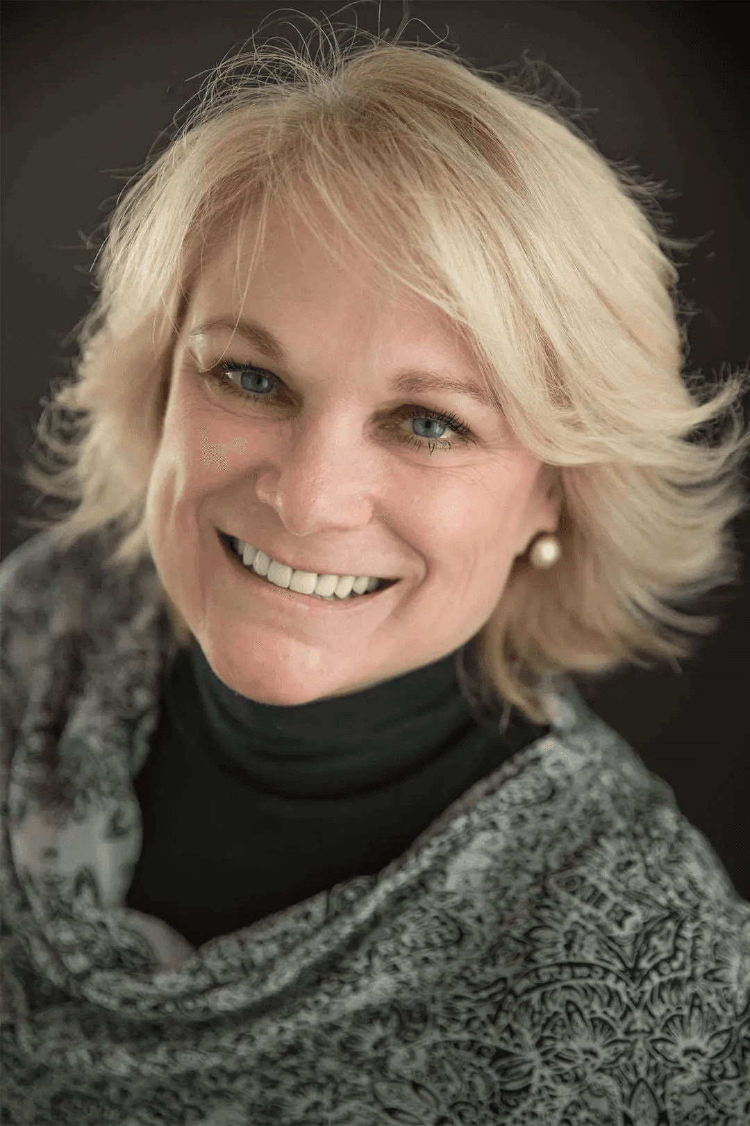A smiling woman with blonde hair wearing a patterned scarf and black top against a dark background.
