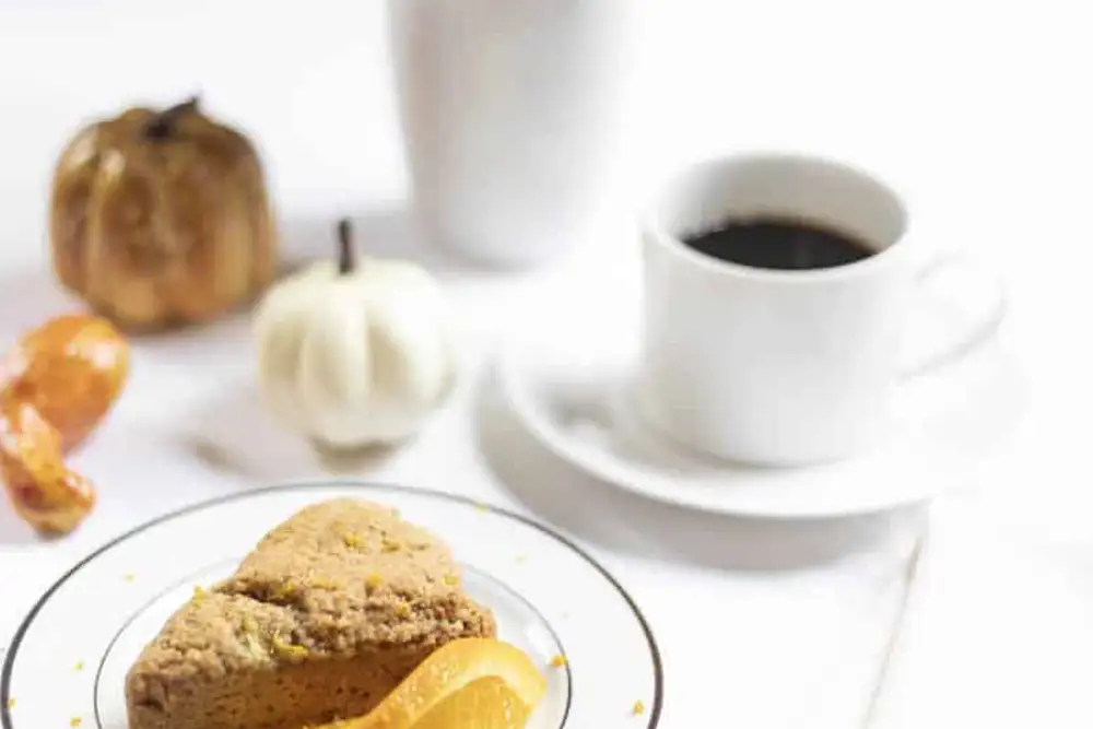 A plate with a scone and orange slice beside a cup of coffee and decorative pumpkins.