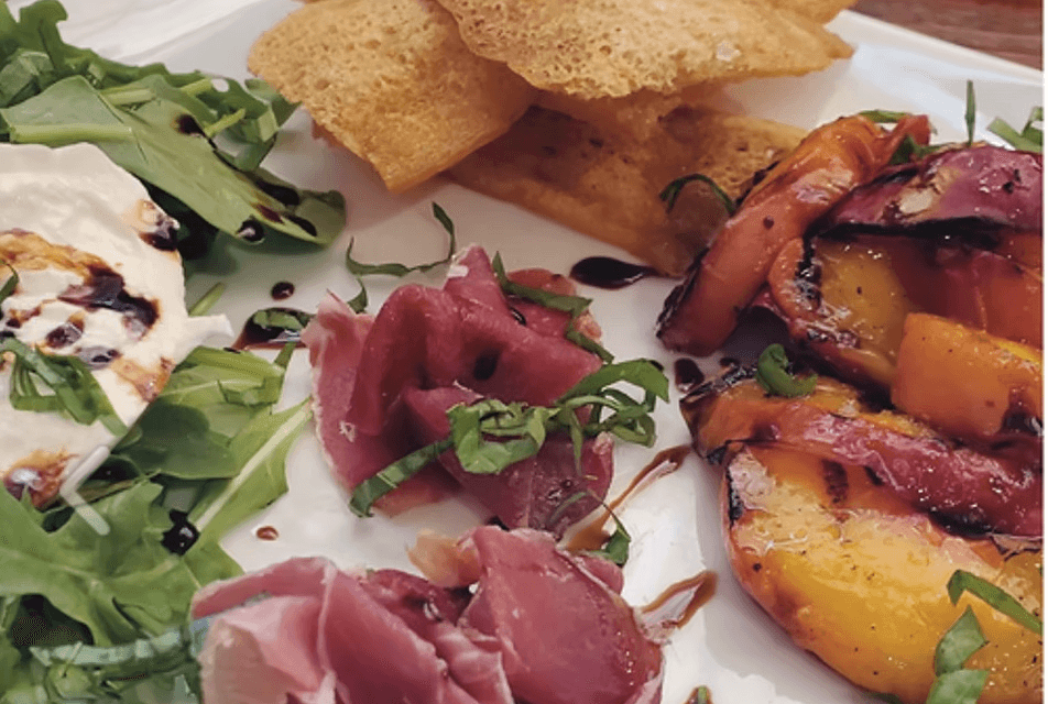 A white square plate with an array of curled up prosciutto, artisan crackers, grilled stone fruit and arugula with balsamic