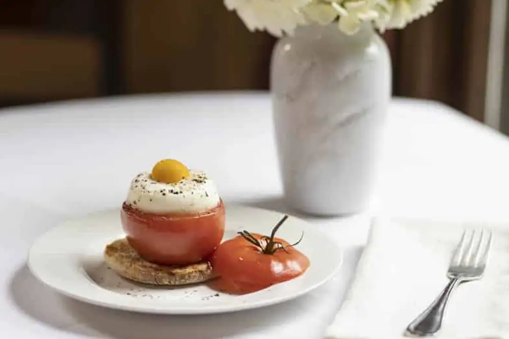 A stuffed tomato topped with cream and an egg yolk, served on a plate with a fresh tomato and a vase of flowers in the background.