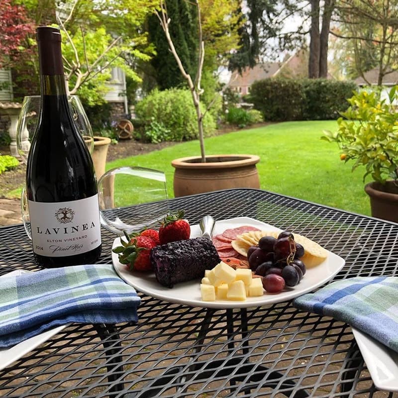 A wine bottle with a plate of assorted cheese, meats, and fruits on a table set in a garden.