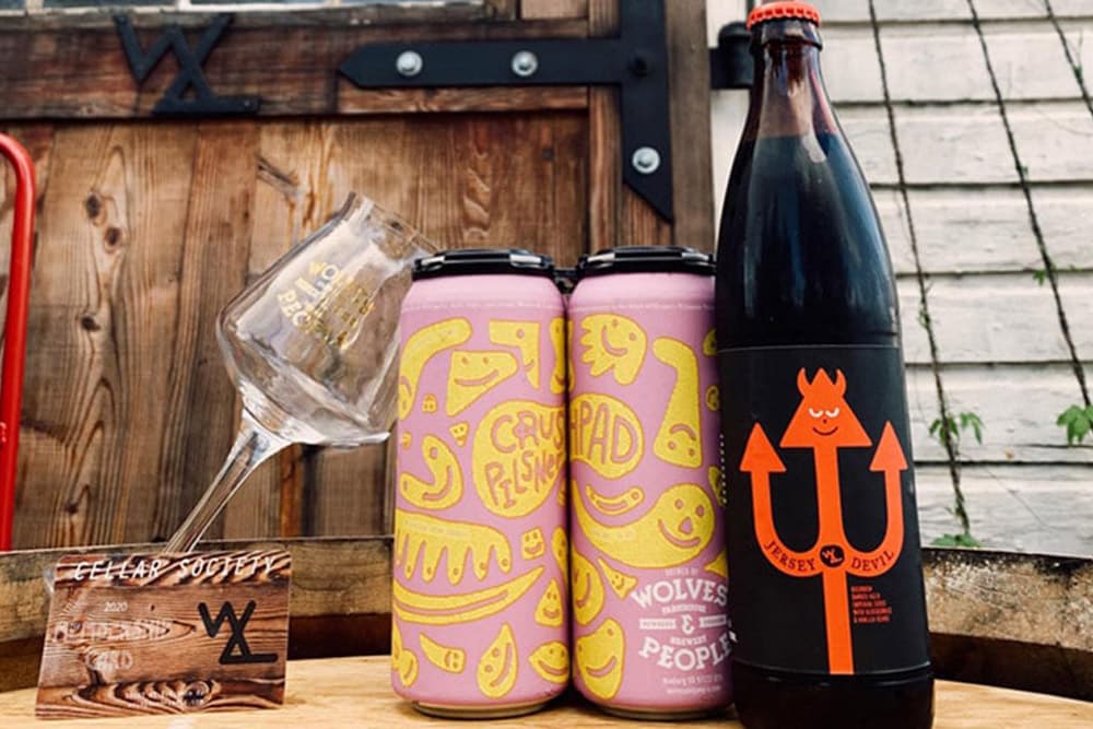 A glass, two colorful beer cans, and a bottle are arranged on a wooden surface with a rustic background.