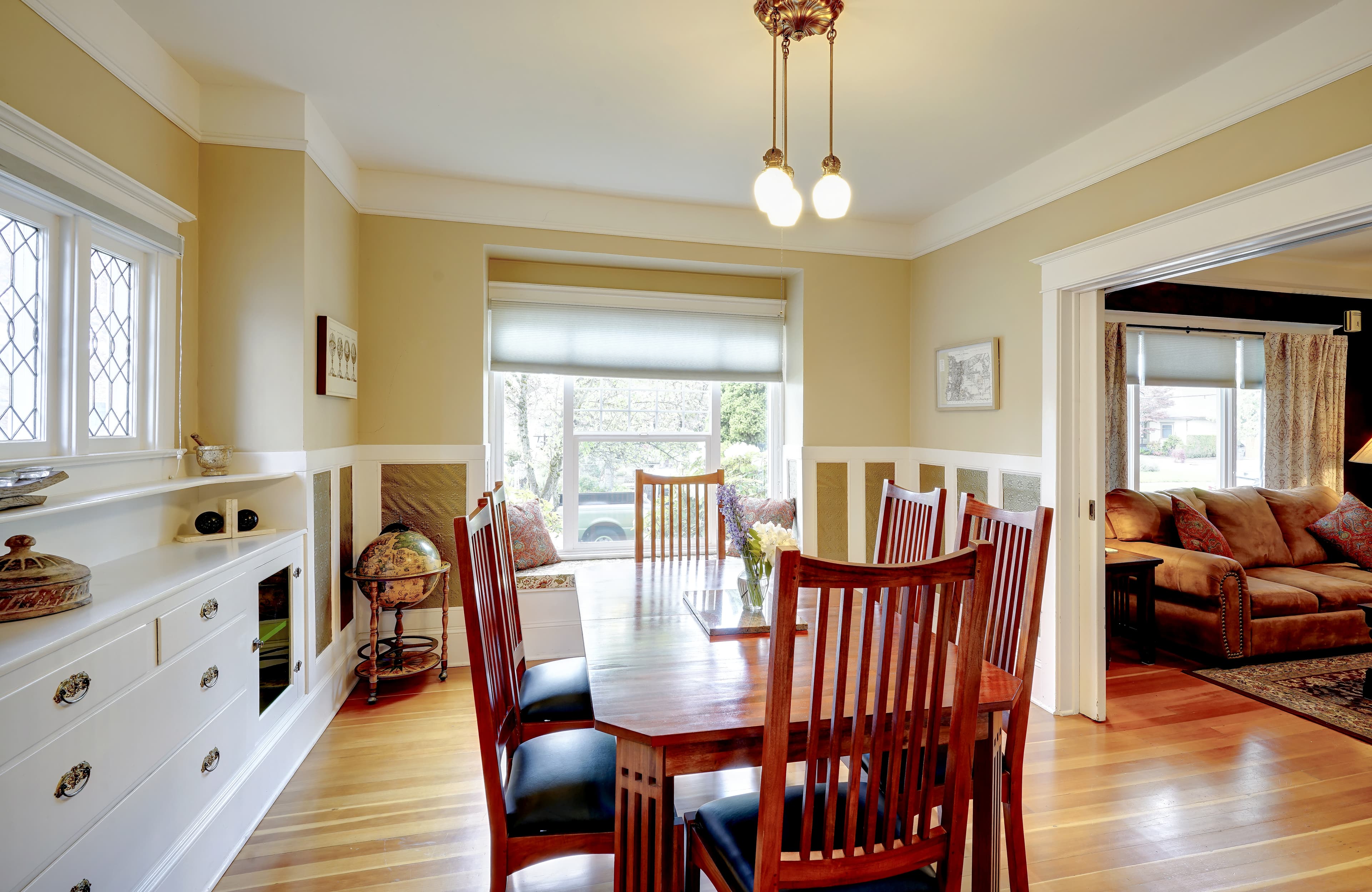 View of dining room from the kitchen with a table surrounded by 6 chairs and a window seat. Hard wood floors.