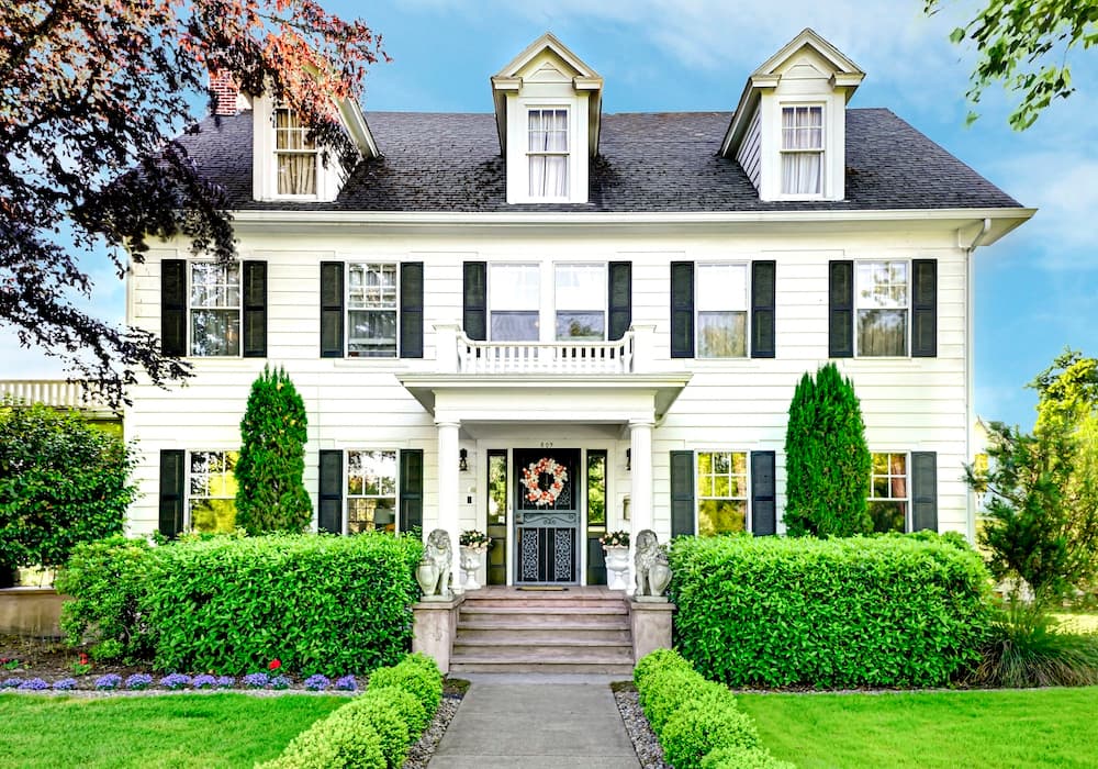 A charming white house with green shutters, flanked by manicured shrubs and a flower garden.