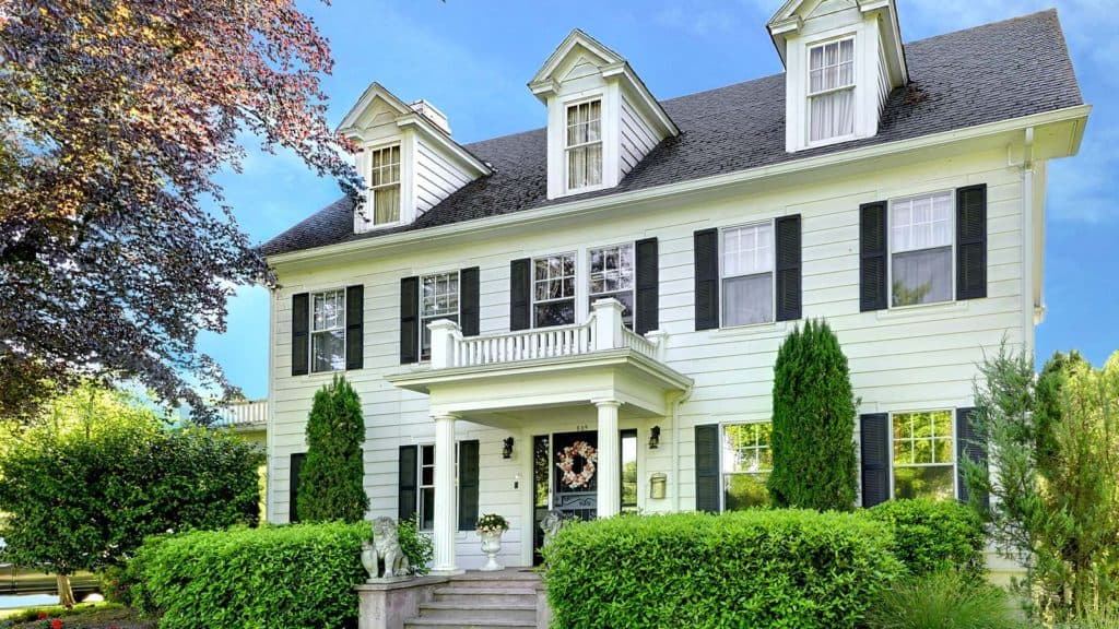 Victorian-style white house with black shutters and a welcoming front porch surrounded by greenery.