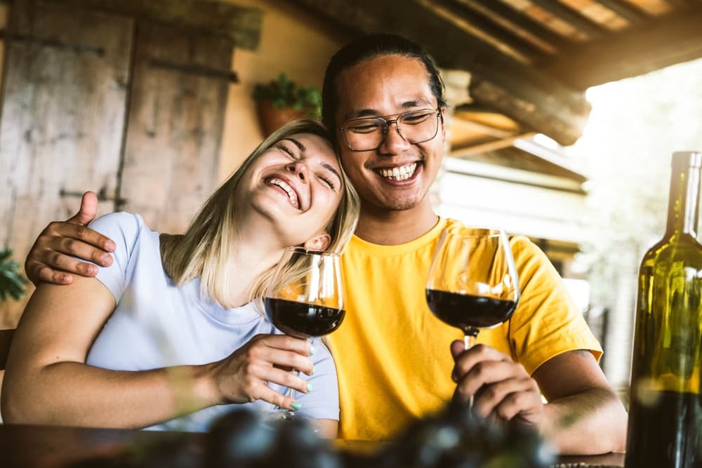 A happy couple smiles while holding glasses of red wine at a cozy table.