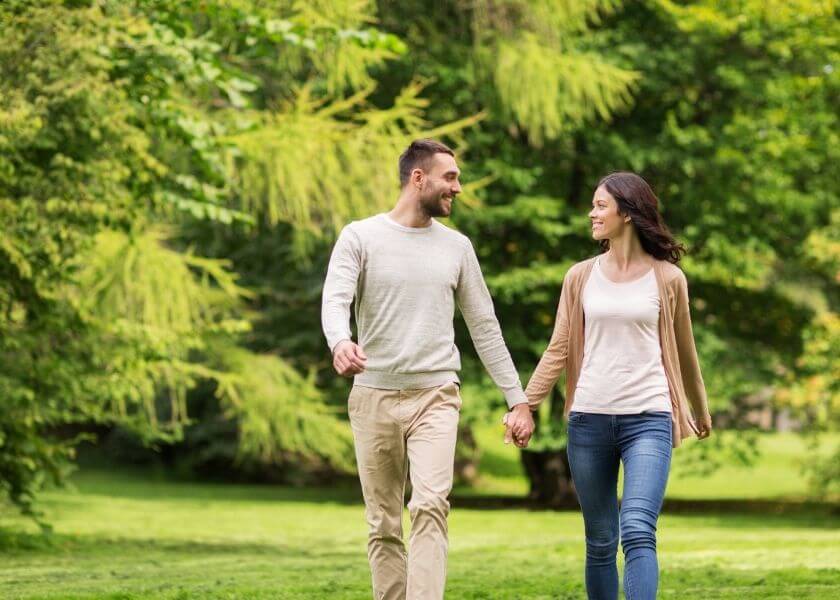 smiling couple walking with hands clasped through a lush park during summer