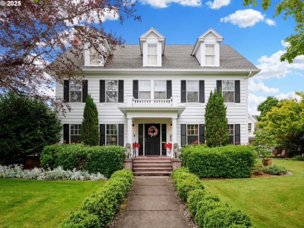 Exterior view of A' Tuscan Estate Bed and Breakfast, a white three-story historic home with black shutters, dormer windows, a manicured lawn, lush greenery, and a welcoming front porch decorated with a wreath and flower pots under a bright blue sky. Exterior view of A' Tuscan Estate Bed and Breakfast, a white three-story historic home with black shutters, dormer windows, a manicured lawn, lush greenery, and a welcoming front porch decorated with a wreath and flower pots under a bright blue sky.