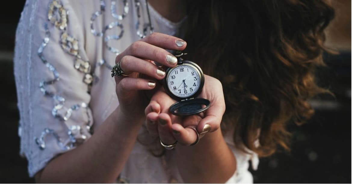 Close-up of a woman holding an antique pocket watch in both hands, wearing a sequined blouse and metallic nail polish, with long wavy hair cascading beside her.