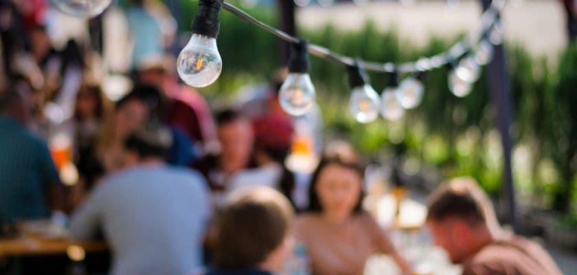 String lights glow above guests blurred in the background as they enjoy a lively evening of patio dining.