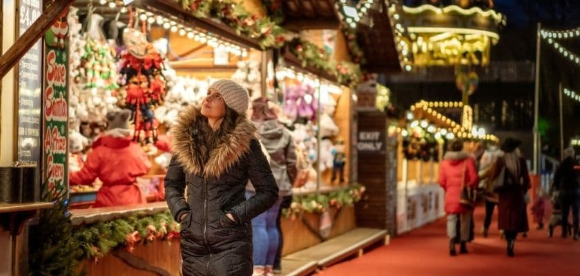 woman in winter coat browses gift stalls at an outdoor christmas market decorated with lights