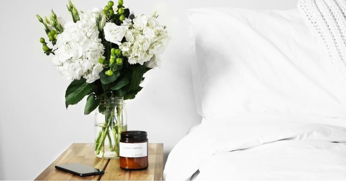 Close-up of a bedside table with a glass jar of white hydrangeas and greenery, an amber candle labeled “Sunday Campfire,” a smartphone, and crisp white bedding with a knit throw.